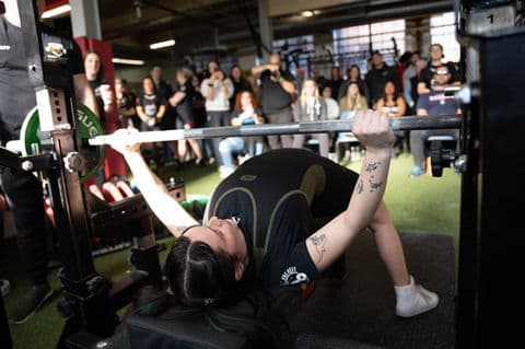 A lifter presses the bar off her chest as spectators watch from the rail.