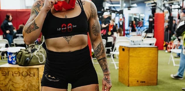 A competitor stands on the warm-up floor between attempts, kit and boxes stacked behind her.
